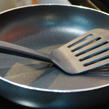 Frying pan holds a black slotted spatula, resting on a stove with a glass cooktop.