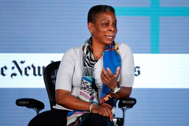 A person in a colorful scarf sits in a chair, gesturing while speaking against a backdrop with text partially reading "The Newâ€¦".
