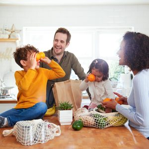 Family interacts playfully with oranges in kitchen, surrounded by groceries and plants, enjoying a bright, sunny day at home.