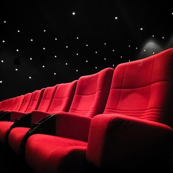 Red cinema seats arranged in a row, illuminated dimly, against a dark theater background with scattered small ceiling lights.