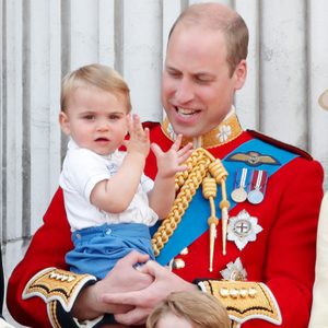 Child waving while being held by an adult in a red ceremonial outfit, adorned with medals and gold braid, against a striped stone background.