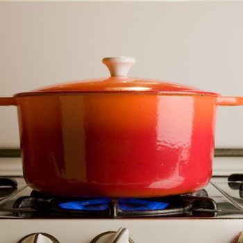 Red pot resting on a stove, covered and heated by a blue flame, in a kitchen setting.
