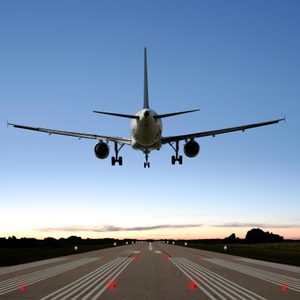Airplane descending onto a runway, illuminated by red landing lights, with a sunset sky and distant trees in the background.
