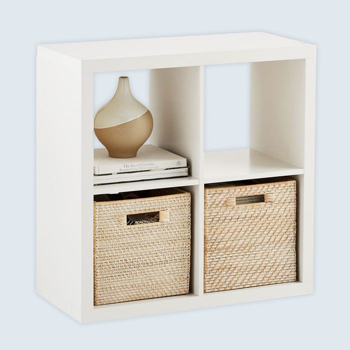 Bookshelf holds vase and books on top shelf, woven baskets below; placed against a plain background.