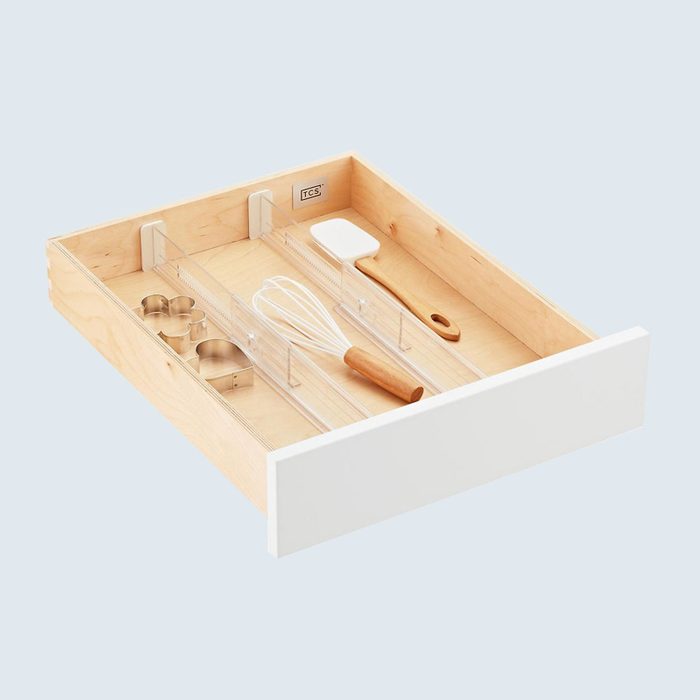 Wooden drawer organizes kitchen utensils, including a whisk, spatula, and cookie cutters, using clear dividers. Set against a light blue background.