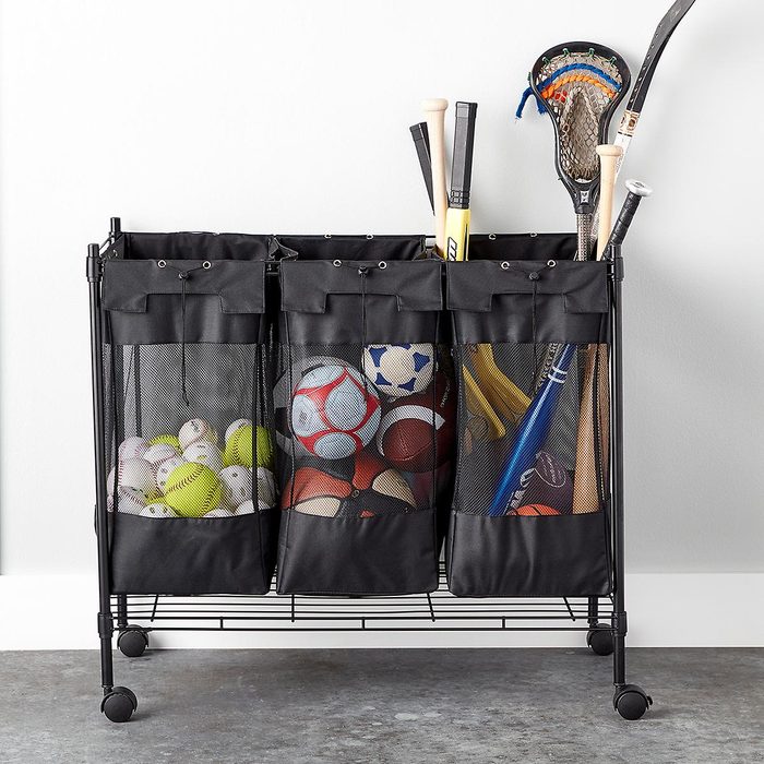 Storage cart holds various sports equipment; baseballs, tennis balls, soccer balls, bats, lacrosse sticks. Positioned against a plain, white wall.