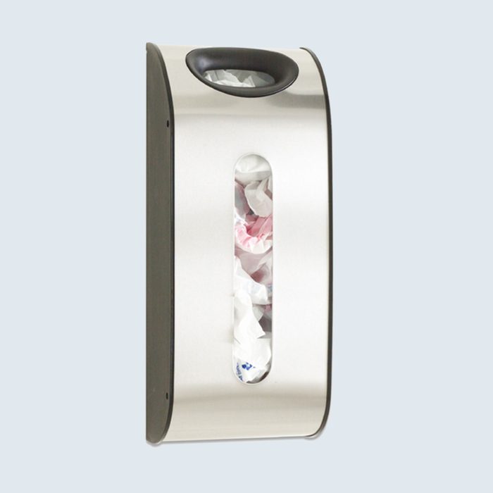 Metal wall-mounted dispenser holds crumpled plastic bags, visible through a vertical window, against a light gray background.