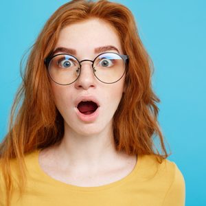 Person with red hair and glasses looks surprised, mouth open, wearing a yellow shirt against a blue background.