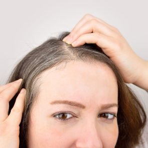 Hands part hair, revealing gray roots, on a person's head with a neutral background.
