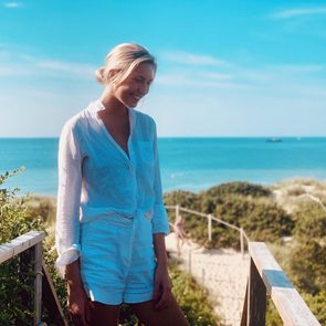 Woman smiling on wooden path overlooking beach, wearing a white outfit, with ocean and sky in the background.