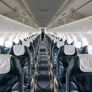 Empty airplane seats align symmetrically, facing forward, inside a brightly lit cabin. Overhead bins are closed above the seats.
