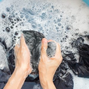 Hands vigorously scrubbing black fabric in soapy water, within a blue basin.