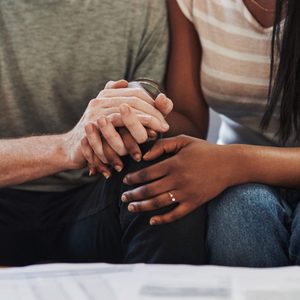 Hands clasp together supportively; two people sit closely on a sofa, with documents visible on a table in front.