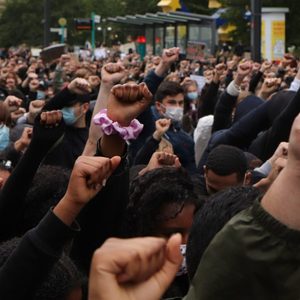 Protesters raise fists in unison, standing closely packed in an outdoor urban area, with some wearing masks amidst a large crowd.