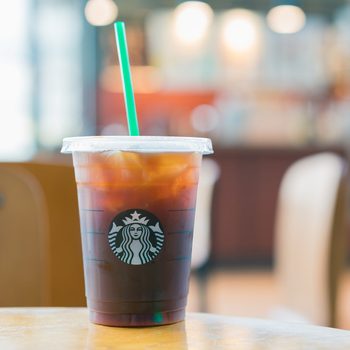 A clear plastic cup of iced coffee with a green straw sits on a table in a blurred cafe background.