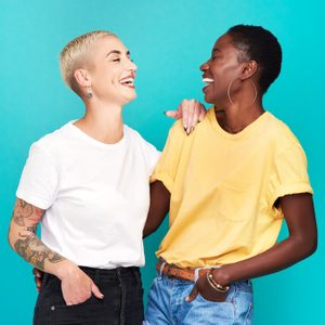 Two people laughing, one with a tattooed arm, the other wearing hoop earrings, stand against a bright turquoise background.