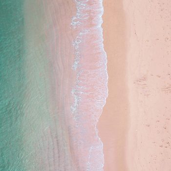 Waves lightly wash onto a sandy beach, creating a contrast between turquoise water and pinkish sand with scattered footprints.