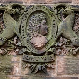 Carved stone relief portraying Elihu Yale's profile, flanked by two goats, set in a brick wall. Text reads: 