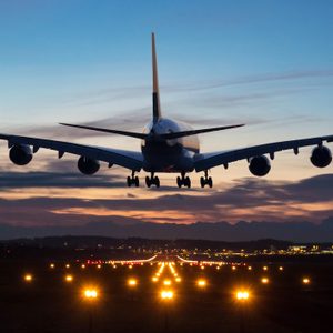 Airplane descends towards illuminated runway during sunset, silhouetted against a colorful sky with distant mountains and city lights in the background.