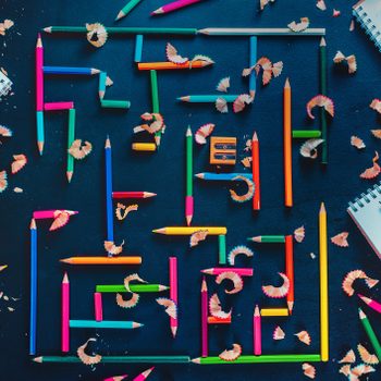Colored pencils form a maze on a dark surface, surrounded by shavings, notebooks, and a sharpener.