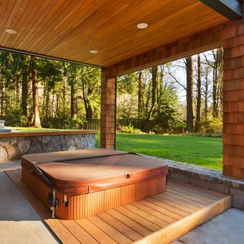 Hot tub sits unused under a wooden patio, bordered by grass and trees in a serene outdoor setting.