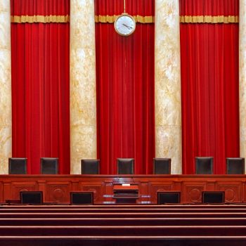 Chairs line a wooden bench, facing rows of pews, surrounded by tall red curtains and marble columns in a formal, courtroom setting.