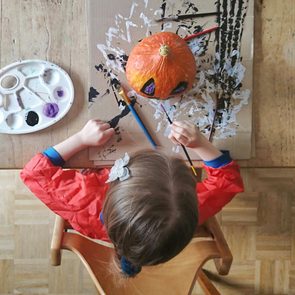 Child paints a pumpkin on a cardboard-covered table, using a palette with various colors.