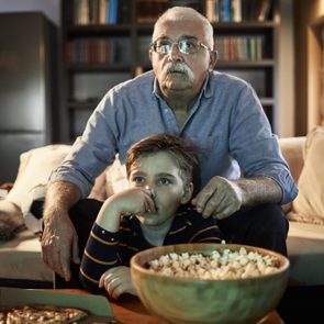 Grandfather and child watch TV at home, seated on a couch, surrounded by popcorn and pizza in a cozy living room with bookshelves.