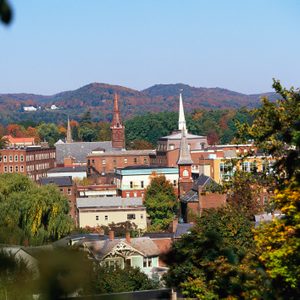 Buildings with spires stand among trees, framed by vibrant autumn foliage, with distant rolling hills under a clear blue sky.
