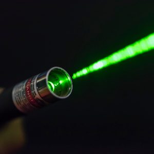Laser pointer emits green beam within a dark setting, held by a hand. Warning label reads 