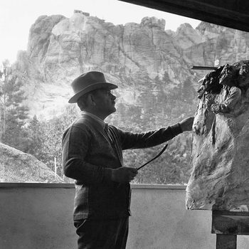 Man in a hat sculpts a bust, standing indoors. Large rocky landscape visible through the window behind him.