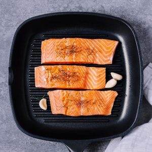 Salmon fillets lie seasoned with herbs in a black grill pan atop a gray surface, accompanied by garlic cloves.