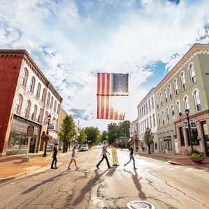 Four people walk across a street beneath a large American flag. Historical buildings line both sides under a partly cloudy sky.