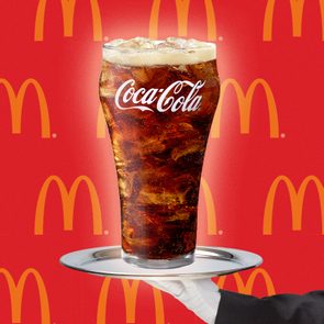 A glass of Coca-Cola with ice rests on a tray held by a hand against a red background with McDonald's logos.