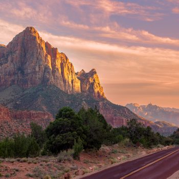 Sunlit mountains rise dramatically, basking under a vibrant sky, adjacent to a winding road and lush greenery.