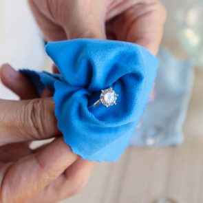 A diamond ring being polished with a blue cloth held by hands on a wooden surface.