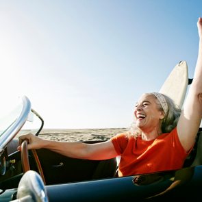 Woman joyfully driving convertible, raising hand. The beach and a clear blue sky form the backdrop. Surfboard visible in the car.