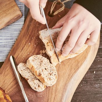Hands slice bread on a wooden board, surrounded by scattered crumbs and cutlery on a table with a blue cloth and a pastry nearby.