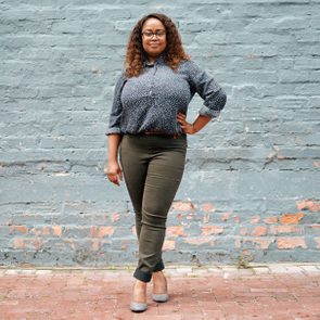 Person stands confidently, hand on hip, against a textured blue brick wall, wearing a patterned shirt, dark green pants, and heels.