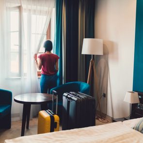 Person gazes out window, standing in a hotel room with two chairs, a table, suitcases, and a floor lamp.