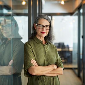 A woman stands confidently with arms crossed in a modern office hallway, reflected in glass panels.