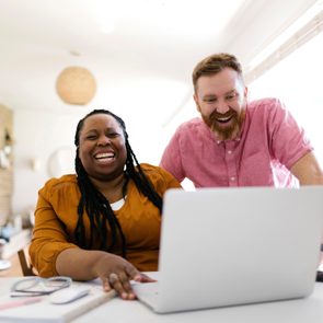 Two people laugh together while looking at a laptop in a bright, modern living room.