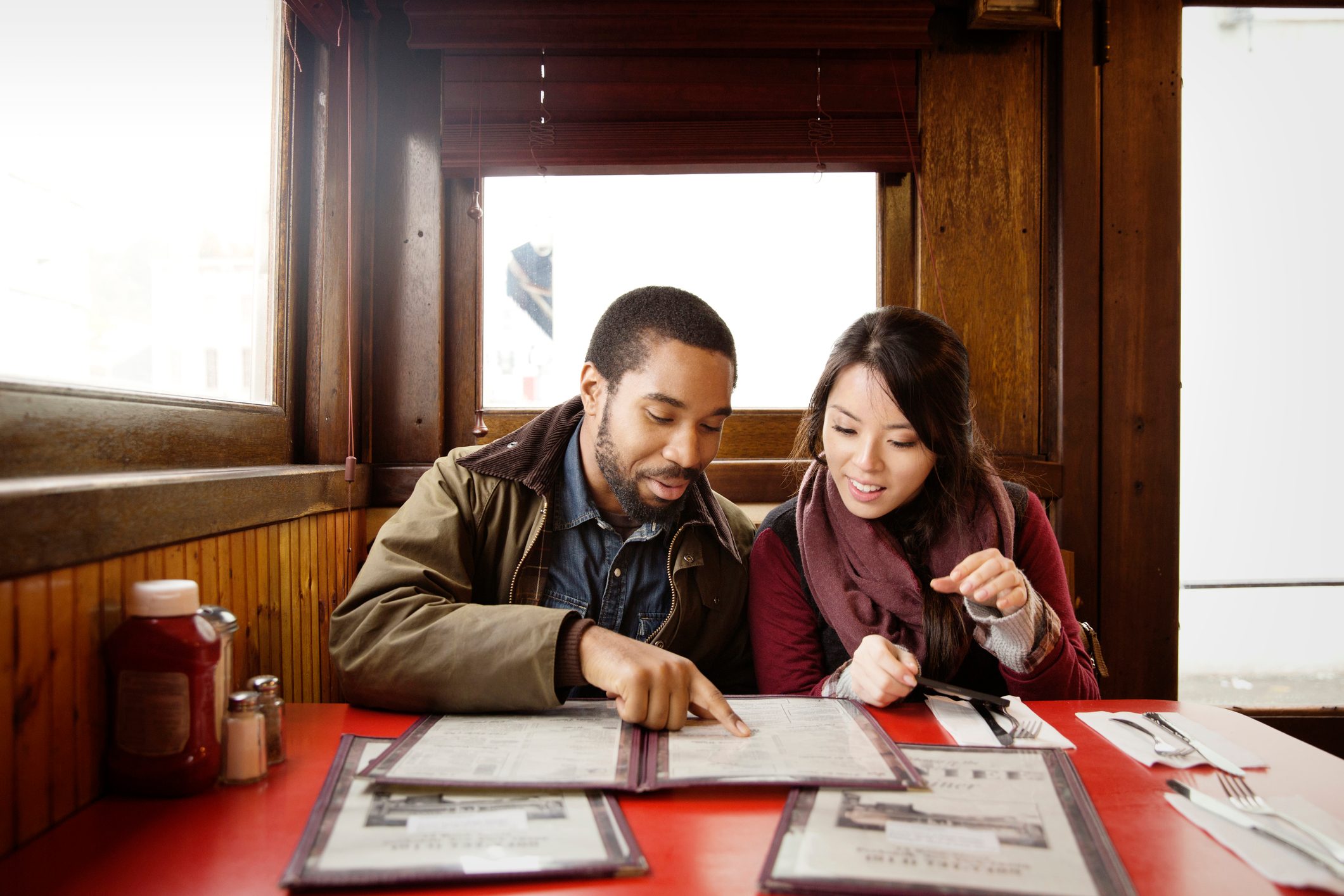 Two people review menus at a red table in a cozy wooden restaurant booth. Ketchup and utensils are placed nearby.