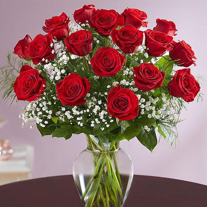 Red roses arranged in a clear vase, surrounded by delicate white flowers and greenery, placed on a dark table against a soft purple wall.