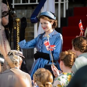 A woman in a blue outfit waves to a crowd holding flags, standing on steps with red carpet, surrounded by admirers.