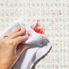 Hand wipes red stain with white cloth on textured light carpet.