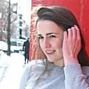 A person smiles, touching their hair, standing against a red wall on a snowy street.