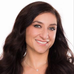 A woman smiles warmly, showing her teeth, with long dark hair, wearing a necklace, against a plain white background.
