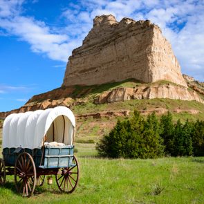 A covered wagon stands idle on green grass, set against a large, rugged bluff under a blue, partly cloudy sky.