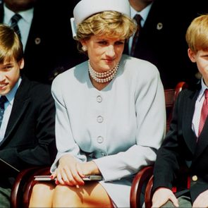 Three people sit on chairs outdoors; a woman in a light outfit between two boys in suits, under sunny conditions.